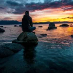Ihminen istuu kivellä järven rannalla auringonlaskussa