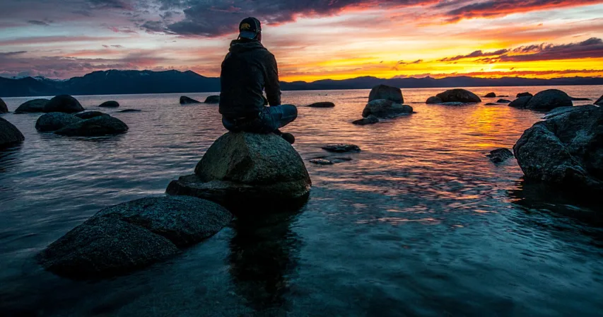 Ihminen istuu kivellä järven rannalla auringonlaskussa
